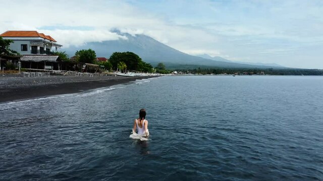 Woman walk in water in white dress with volcano view near to black sand beach in Amed, Bali, Indonesia. High quality 4k aerial footage