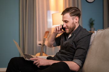 Smiling man is sitting in the living room on the sofa with his laptop on his legs, browsing vacation deals, hotels, looking for the best bargains, calling on the phone and booking a trip, holiday