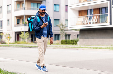 Fototapeta premium food shipping, profession and people concept - happy smiling indian delivery man with thermal insulated bag and smartphone walking along city street