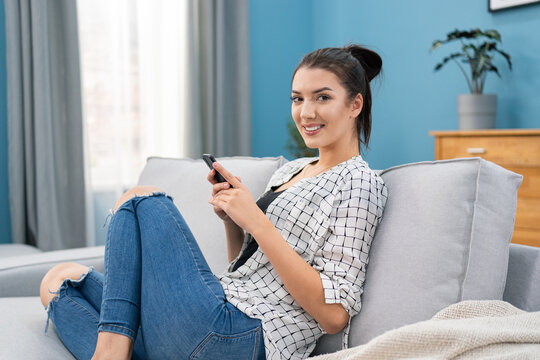 Influencer sits on sofa in living room holding phone in hands, she sends messages to her friends, writes a blog post, checks the number of likes, followers, replies to comments under a new photo