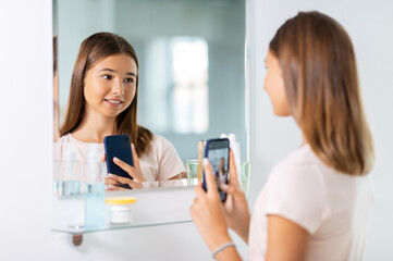 beauty, hygiene and people concept - teenage girl looking in mirror and taking selfie with smartphone at bathroom