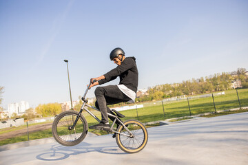 Crazy cyclist rides his low bike at the skatepark, bmx lifting the front wheel and being in the air twists his body, hips, performs tricks, laughs, smiles in the background trees, nature - obrazy, fototapety, plakaty