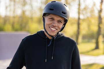 A dark-skinned man wearing a helmet standing in a park resting after a bike ride, smiling, his...