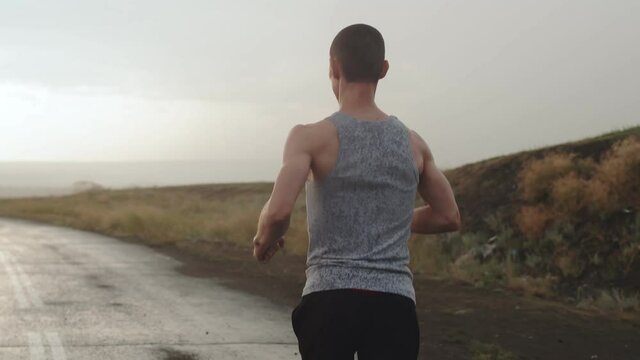 Man of athletic build runs along road in countryside during the rain, flash of lightning, view back. Runner continues his run during thunderstorm. Concept of perseverance