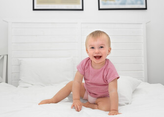 Fototapeta premium Happy caucasian baby girl about 1 year old having her two first milk teeth laughing sitting on bed.Adorable child playing indoors,kid having fun before going to bed, after waking up or toothing idea