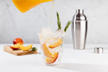 Cooking healthy summer peach juice - cocktail pour of jug in glass with ice, rosemary twig, sugar rim, ingredients in elegant white interior on soft light wood table.