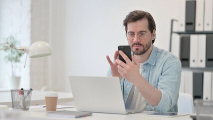 Young Man with Laptop using Smartphone at Work