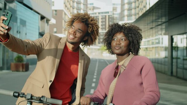 Young positive Afro-American man and woman standing with electric scooters in city and smiling at smartphone camera while taking selfie