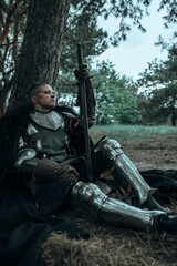 Medieval knight in armour sits and rests in forest with sword in his hand.