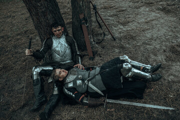 Two tired medieval knights rest among forest under tree.
