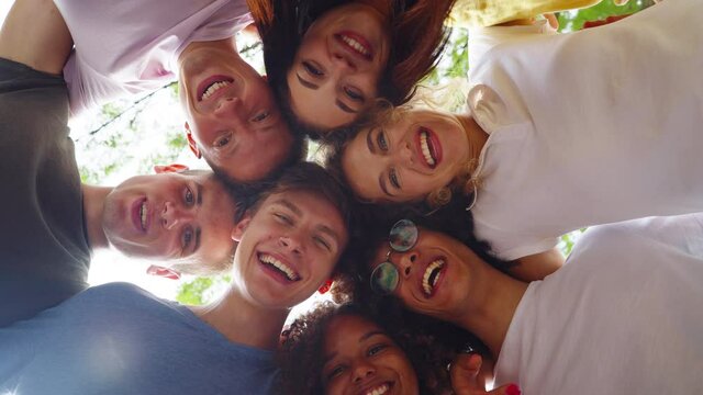 Young multiethnic people in casual wear bending above camera and smiling, green trees and sky on background. Low angle happy friends together. Concept of bonding