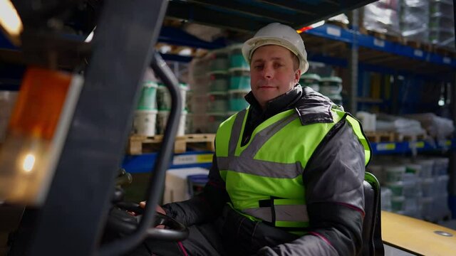 Portrait of confident Caucasian man in hard hat and vest sitting in forklift in warehouse looking at camera smiling. Professional loader posing at workplace indoors. Industrial concept