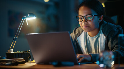 Young Teenage Multiethnic Black Girl Using Laptop Computer in a Dark Cozy Room at Home. She's Browsing Educational Research Online. Studying Science School Homework Concept.