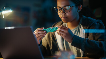 Young Teenage Multiethnic Girl Using Laptop Computer and Looking at Printed Circuit Board in Cozy Room at Home. She's Browsing Educational Research Online. Studying Science School Homework Concept.