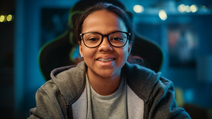 Portrait of a Cute Young Teenage Multiethnic Black Girl in a Dark Cozy Room at Home. She's Happy, Looks at Camera and Smiles. Young Female Wears Glasses and Braces.