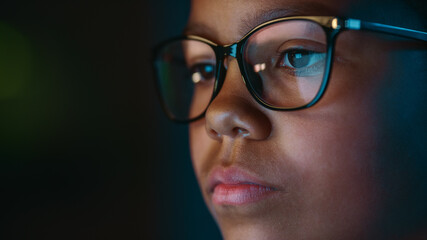 Fototapeta na wymiar Close Up Portrait of a Cute Young Teenage Multiethnic Black Girl in a Dark Cozy Room at Home. Young Female Wears Glasses that Reflect School Homework from Her Laptop Computer Screen in Them.