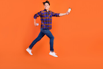 Full size profile photo of millennial guy jump hold coffee telephone wear shirt jeans sneakers isolated on orange background