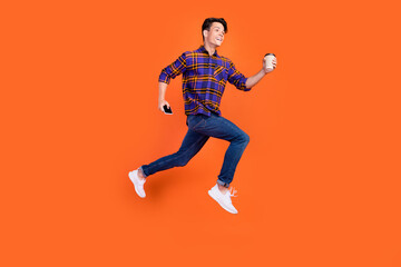 Obraz na płótnie Canvas Photo of hurrying teenager man dressed checkered clothes jumping running holding coffee phone isolated orange color background