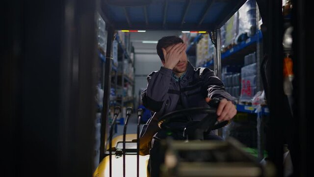 Tired young handsome forklift driver taking off white hard hat wiping forehead sitting in vehicle in warehouse. Portrait of exhausted confident Caucasian man working in logistics industry