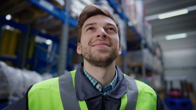 Close-up portrait of handsome brunette unshaved man with brown eyes smiling looking at cargo racks in industrial storage. Headshot of satisfied Caucasian male worker in warehouse indoors