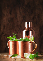 Moscow mule cocktails in copper mug with lime, ice, ginger beer, vodka and mint. Wooden background, bar tools, negative space