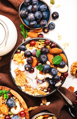 Muesli bowl and organic ingredients for healthy breakfast. Granola, nuts, blueberry, cranberry, oatmeal, greek yoghurt, whole grain flakes on white table