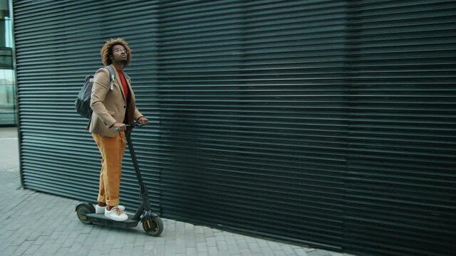 Young Afro-American man in smart casual outfit riding electric scooter in city
