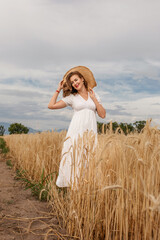 a young attractive woman in a white dress and a straw hat on a field of ripe cereals against a blue sky with clouds in autumn, the concept of harvesting, agribusiness and agriculture