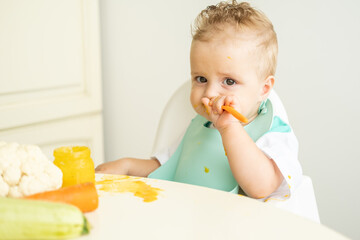 funny baby boy in bib eating vegetable puree with spoon sitting in child chair. Child learn to eat by himself