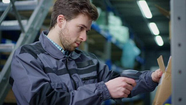 Brunette man scanning barcode on cardboard box with scanner in urban industrial warehouse. Portrait of concentrated handsome Caucasian employee checking stock in storage or terminal