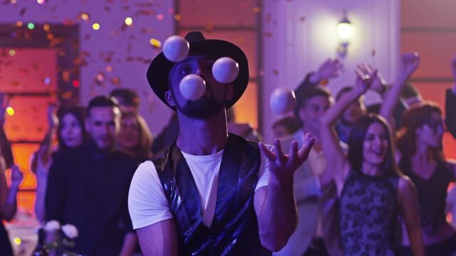 Juggler showing tricks with white balls at colorful party . Man juggling four balls at night party. Many out of focused people in the background . Shot on RED DRAGON Cinema Camera in slow motion.