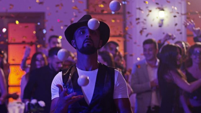 Juggler showing tricks with white balls at colorful party . Man juggling four balls at night party. Many out of focused people in the background . Shot on RED DRAGON Cinema Camera in slow motion.