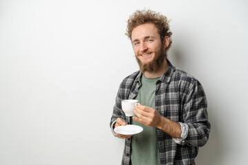 Smiling handsome man with curly hair and beard drinking coffee holding cup, wearing plaid long sleeve shirt isolated on white background. 