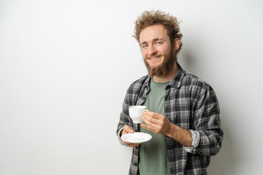 Smiling handsome man with curly hair and beard drinking coffee holding cup, wearing plaid long sleeve shirt isolated on white background. 