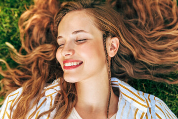 Fototapeta premium Closeup portrait of a attractive young woman looking directly to the camera lying on the green grass with long red hair outdoors.