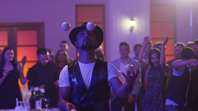 Juggler showing tricks with white balls at colorful party . Man juggling four balls at night party. Many out of focused people in the background . Shot on RED DRAGON Cinema Camera in slow motion.