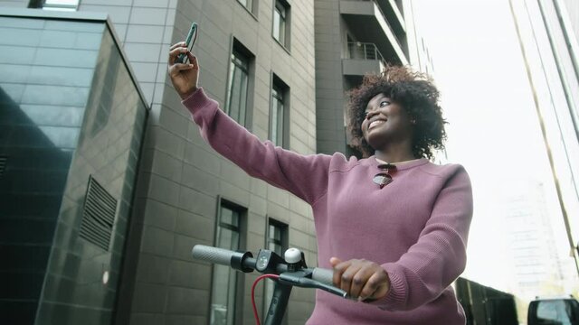 Low angle shot of young smiling Afro-American woman standing with e-scooter in city downtown, taking selfie and then checking pictures on smartphone