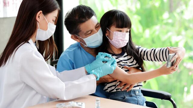 Asian  senior doctor wearing gloves and isolation mask is making a COVID-19 vaccination in the shoulder of child patient with her father at hospital.closed up photo.