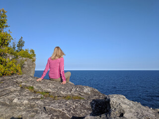 Young woman sits on a high rocky cliff edge overlooking the sea on a sunny day. Bruce Peninsula National Park, Georgian Bay, Ontario, Canada. Hiking, camping, active lifestyle concept.