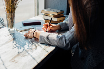 A young woman is sitting at a table by the window and writing in a notebook. The sun's rays fall on...