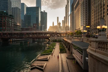 Tuinposter Beautiful downtown Chicago morning along the river as people jog on the path below and train crosses a bridge as the sun casts yellow light into the scene from behind the high-rise buildings beyond. © Joseph Kirsch