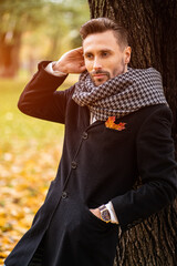 Standing leaned on the tree handsome man straightens his hair wearing dark blue coat. Young freelancer man stands on the street in an autumn coat looking happy waiting foe a date.