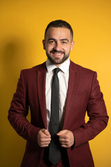 Young handsome man wearing amazing elegant burgundy suit looking on you smiling with hands holding a jacket isolated on yellow background. Men fashion concept. 