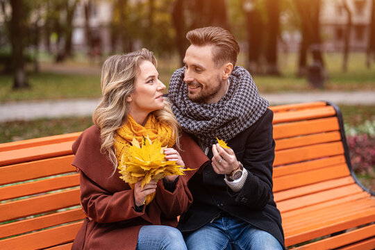 Embracing man found a small leaf happy couple sitting on the bench hugged in park wearing coats and scarfs collecting a bouquet of fallen leaves. Love story concept. Tinted image. 