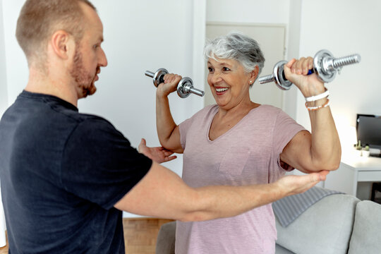 Happy beautiful senior woman smiling and holding easy bar while exercising with personal trainer. An older woman lifting weights in rehabilitation center. Elderly lady is exercising with man at home