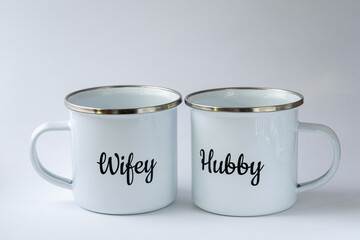 Two white enamel mugs with writings "Hubby" and "Wifey"on them on a white background. Wedding accessories, cups for coffee or tea, luxury wedding.