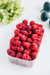 Fresh ripe raspberries in plastic box on white table in kitchen, close-up.
