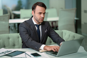 Fototapeta na wymiar Freelancer sitting in front laptop in bright coworking space. Handsome man in business suit working on laptop, freelancer job in progress.
