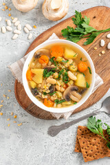 Soup with barley, beans, mushrooms, potatoes, carrots, celery, and herbs. Barley soup with vegetables in a white bowl on a gray concrete background top view. Vegan food, homemade soup.