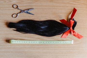 Mechón de pelo cortado anudado con un lazo de raso rojo junto a una cinta métrica y unas tijeras. Donación de cabello para pelucas para pacientes con cáncer - obrazy, fototapety, plakaty